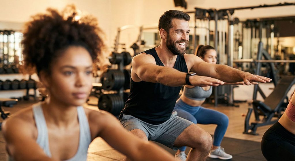A group of people performing squats during a fitness class, focusing on a smiling man in the center.