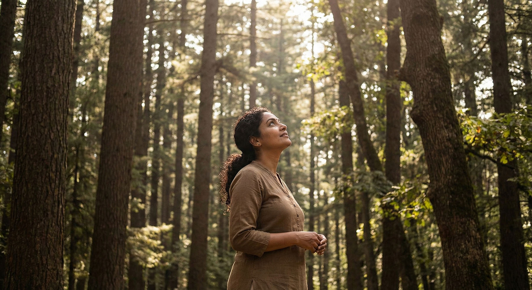 South Asian woman looking up at sunlight filtering through a forest canopy.
