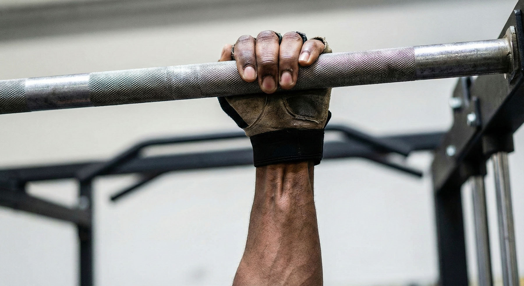 A close-up view of a muscular male forearm and hand, wearing a weightlifting glove, gripping a metal pull-up bar.