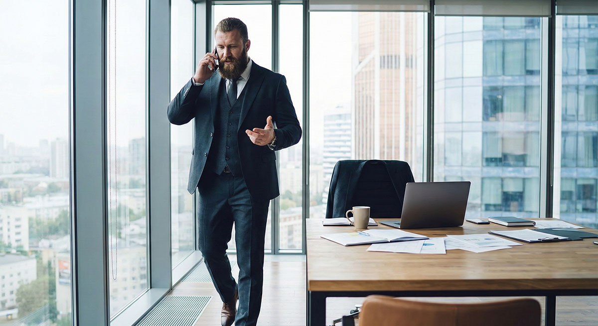 A tall, muscular, bearded businessman in a dark three-piece suit pacing in a modern high-rise office while talking on a cell phone.