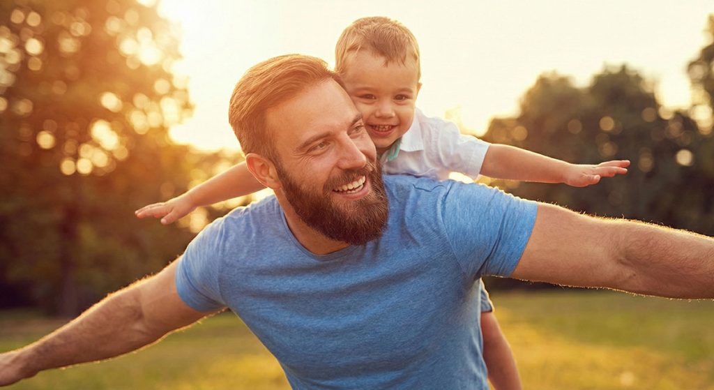 Smiling bearded father giving his young son a piggyback ride in a sun-drenched park.
