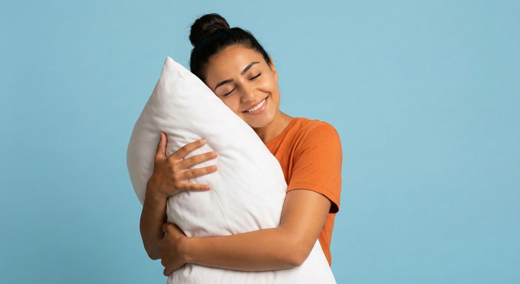 A woman with her hair in a bun smiles peacefully with her eyes closed, tightly hugging a large white pillow against a blue background.