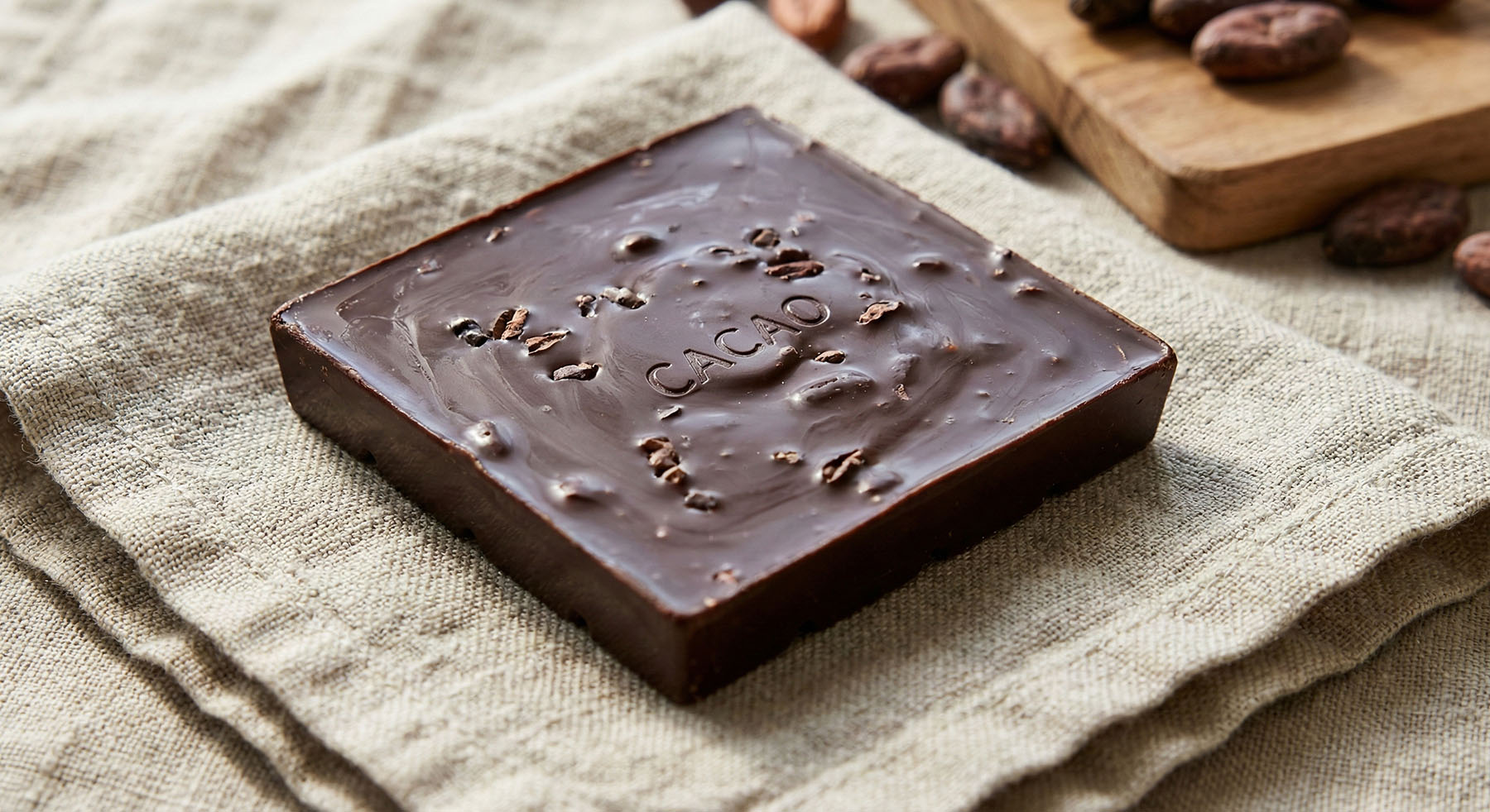 Close-up of a square dark chocolate bar with cacao nibs and "CACAO" stamped on its surface, resting on a linen napkin.