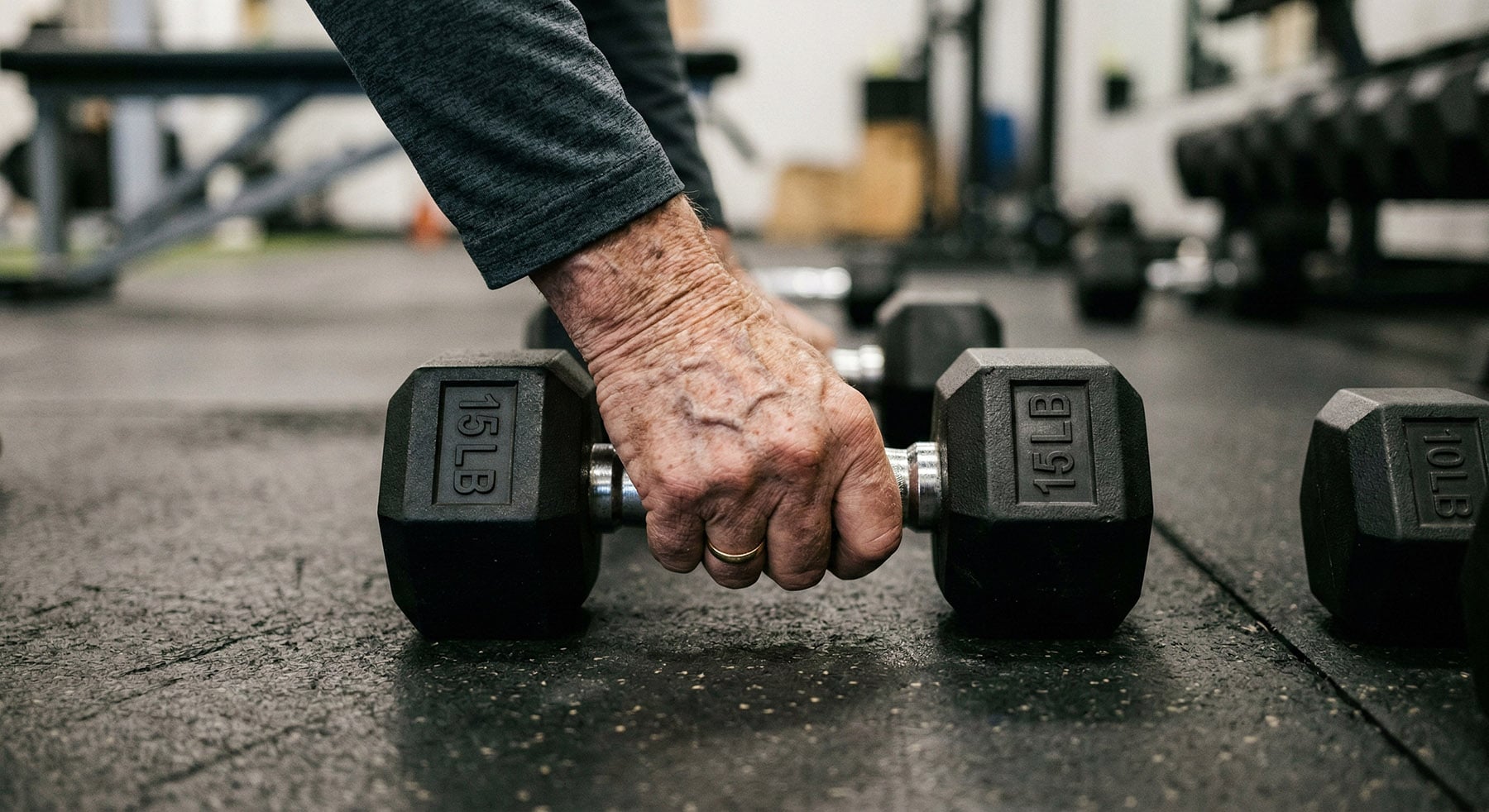 A close-up view of an elderly, wrinkled hand gripping a 15-pound hexagonal dumbbell on a gym floor.