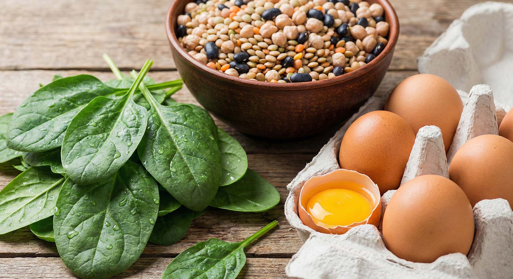 Bowl of mixed lentils, fresh spinach, and a carton of brown eggs with one cracked egg on a wooden table.