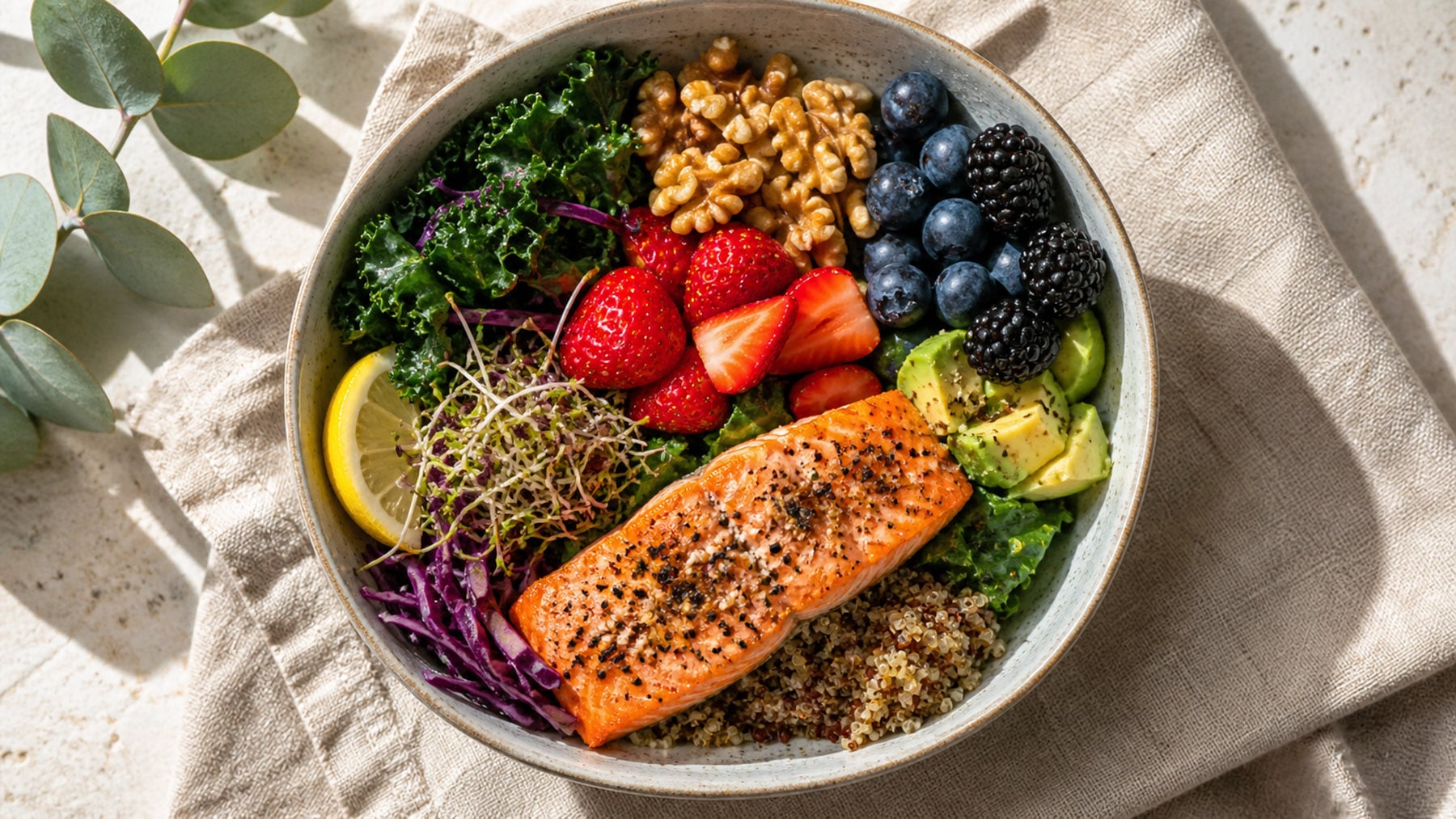 Overhead view of a colorful bowl with salmon, berries, leafy greens, walnuts, quinoa, avocado, cucumber, broccoli, and seeds on a linen napkin.