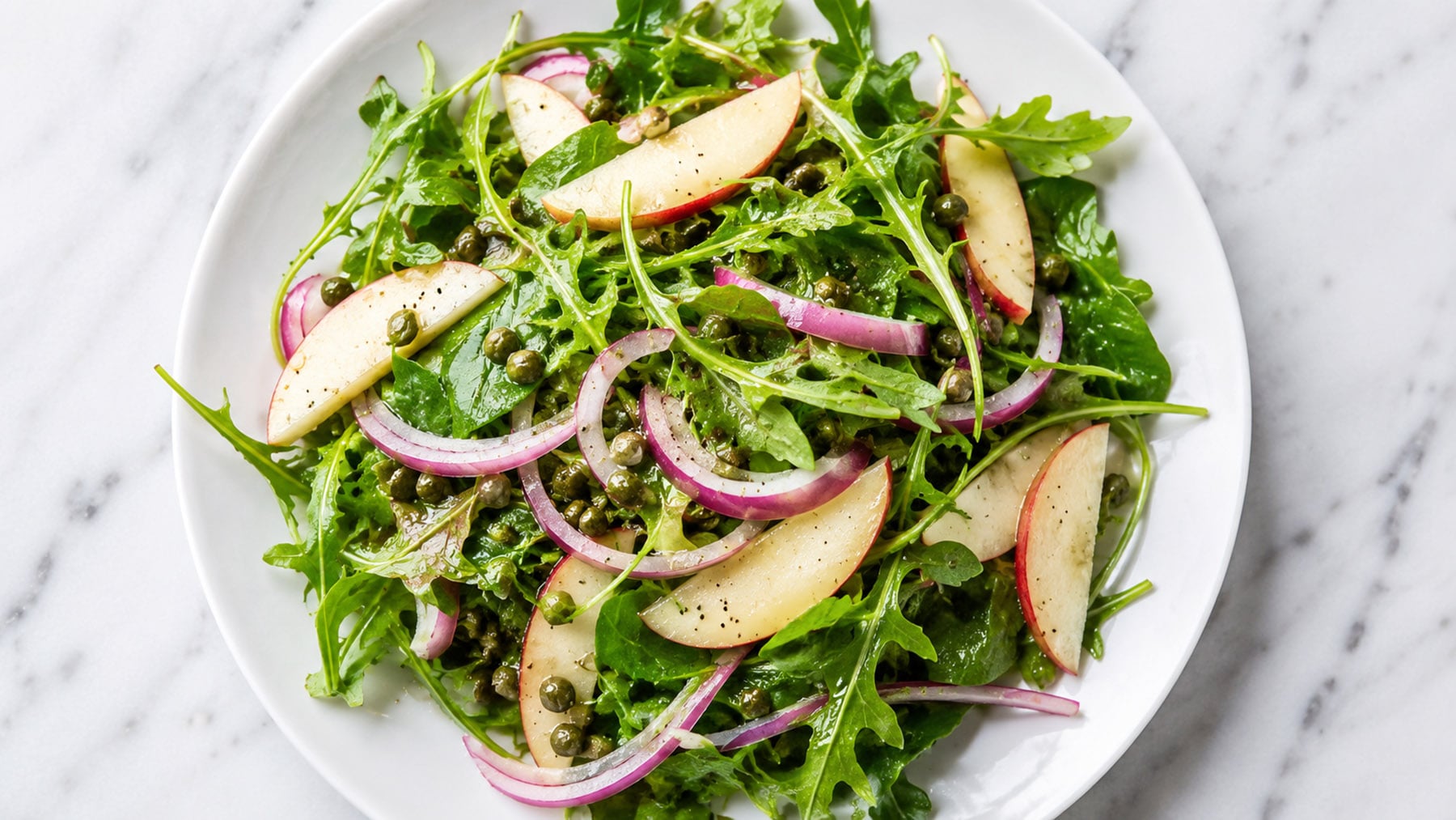 Overhead view of a fresh salad with arugula, red onions, apple slices, and capers on a white plate.