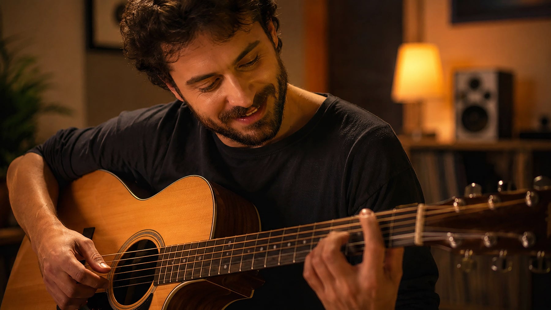 Person smiling while playing an acoustic guitar in a warm, lamp-lit room.