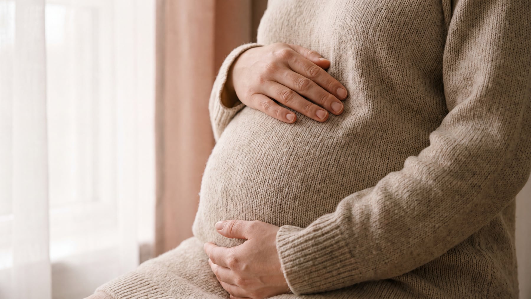 A pregnant woman in an oatmeal sweater cradling her abdomen with both hands beside a sunlit curtain, illustrating Group B strep in pregnancy.