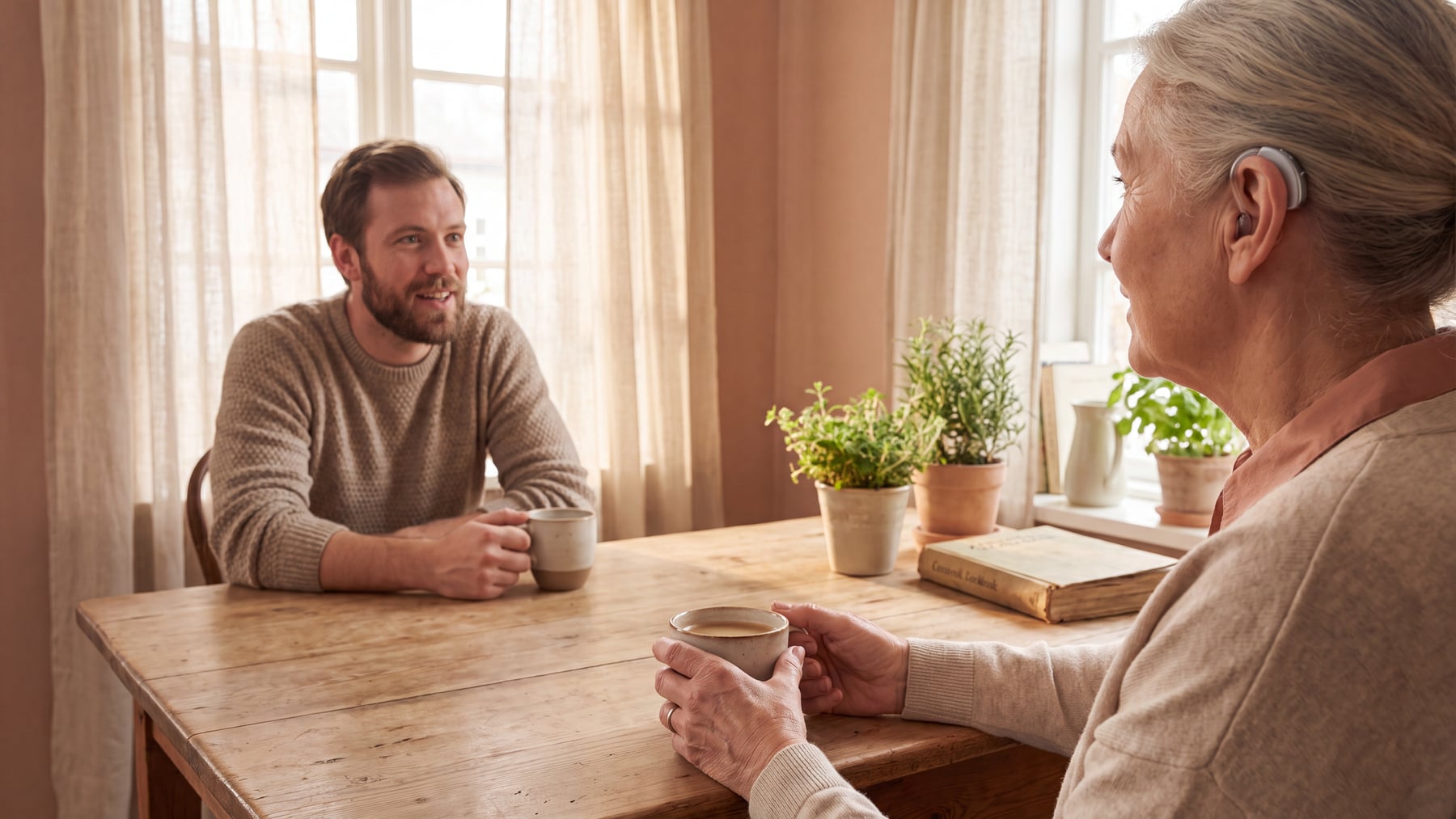 An older woman with a hearing aid in conversation with a younger man across a kitchen table in warm daylight, illustrating the link between hearing loss and dementia.