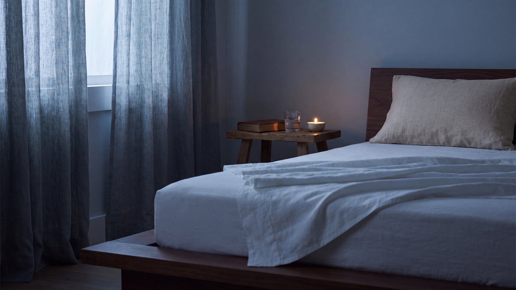 A calm blue-grey bedroom at first light, with white linen bedding and a wooden nightstand holding a lit candle and a glass of water, illustrating the link between sleep and testosterone.