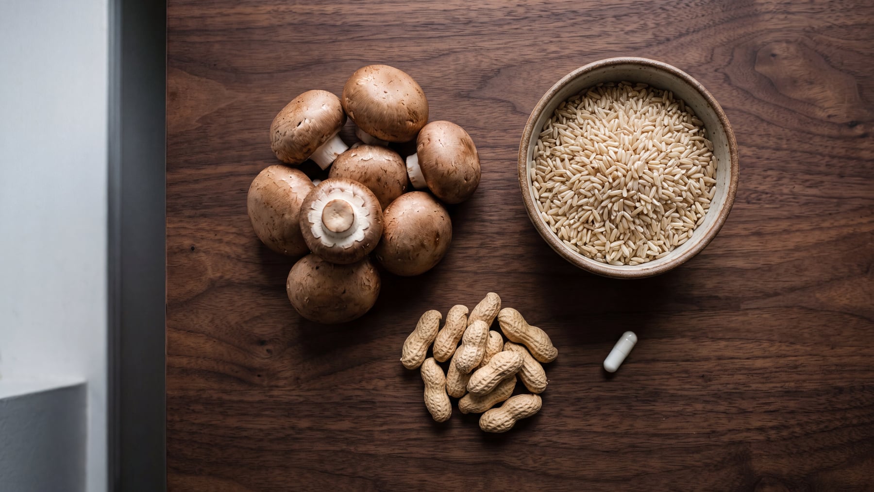 An overhead view of brown mushrooms, peanuts in the shell, a bowl of brown rice and one white capsule on a walnut table, illustrating nicotinamide and DNA repair.