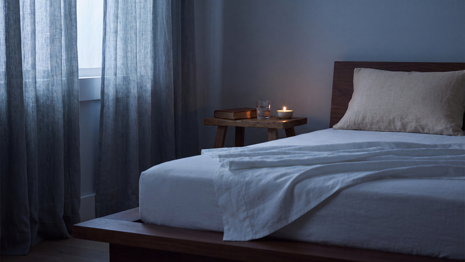 A calm blue-grey bedroom at first light, with white linen bedding and a wooden nightstand holding a lit candle and a glass of water, illustrating the link between sleep and testosterone.